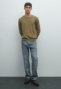 Giovane uomo in piedi contro un muro semplice, indossa un maglione oliva, jeans blu chiaro e scarpe stringate marrone scuro, con le mani dietro la schiena.