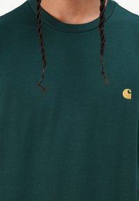 Dark green cotton t-shirt with a ribbed crew neckline, featuring a small yellow logo on the front side. Soft texture, standard fit.