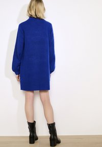 Frau mit blonden Haaren, die ein blaues langärmliges Strickkleid und schwarze, blockabsatz Ankle Boots trägt und vor einer weißen Wand steht.