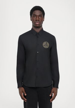Chemise noire à manches longues avec boutons, dotée d'un col, de boutons sur le devant et d'un patch logo doré rond sur la poitrine gauche.