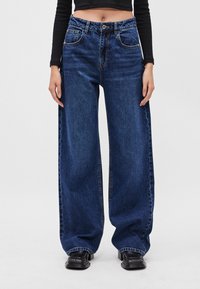 Modré široké nohavice vyrobené z denimu, s vysokým pásom, prednými vreckami a kontrastným šitím. Kombinované s čiernymi robustnými topánkami.