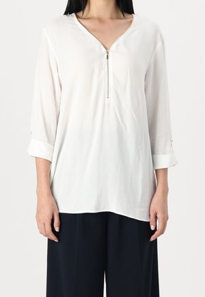 Donna che indossa una blusa bianca larga con una zip frontale e maniche arrotolate, abbinata a pantaloni neri larghi, in piedi davanti a uno sfondo semplice.