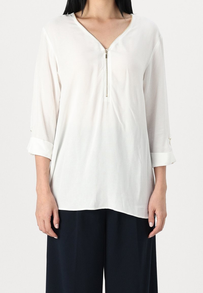 Donna che indossa una blusa bianca larga con una zip frontale e maniche arrotolate, abbinata a pantaloni neri larghi, in piedi davanti a uno sfondo semplice.