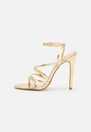Sandale dorée à talons hauts avec lanières, boucle à la cheville, talon fin et design à bout ouvert sur fond blanc.