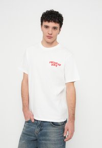 Bílé bavlněné tričko s červeně vyšitým nápisem „FRUITS OF LOVE“ na levé straně hrudi. Volný střih s krátkými rukávy a kulatým výstřihem.