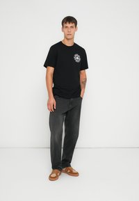 Camiseta negra de manga corta con un gráfico blanco circular en el pecho, combinada con jeans gris oscuro y zapatos marrones con detalles texturizados.