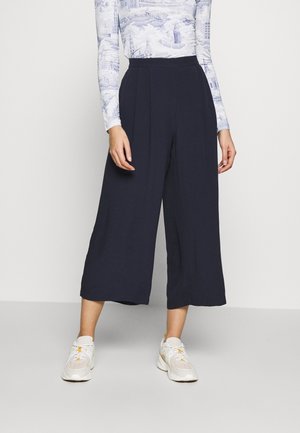 Frau trägt hoch taillierte, weit geschnittene schwarze Cropped-Hosen, weißes langärmeliges Oberteil mit Muster und weiße Sneaker, steht auf hellgrauem Boden.