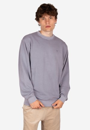 Junger Mann mit lockigem Haar, der einen hellvioletten Pullover und beige Hosen trägt, steht vor einem schlichten weißen Hintergrund.