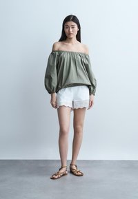 Žena stojaca v zelenej blúzke s odhalenými ramenami, bielych šortkách so zúbkovaným okrajom a hnedých sandáloch s remienkami na jednoduchom svetlom pozadí.
