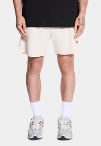 Męski model stoi, ubrany w czarną koszulkę, jasne szorty w kolorze kremowym i szare sneakersy z białymi skarpetkami, prezentując widoczne tatuaże na nogach.