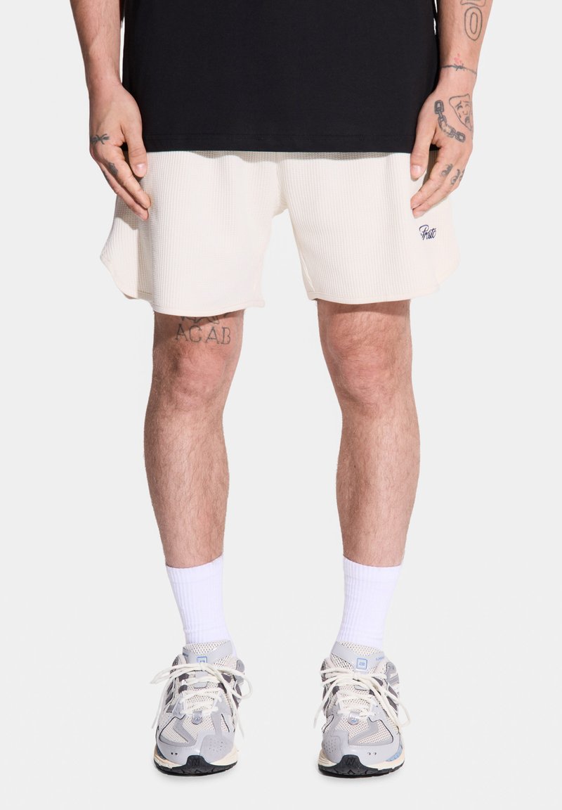 Męski model stoi, ubrany w czarną koszulkę, jasne szorty w kolorze kremowym i szare sneakersy z białymi skarpetkami, prezentując widoczne tatuaże na nogach.