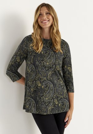 Kvinde med langt bølget hår iført en sort tunika med paisley-mønster og trekvartlange ærmer, smilende og stående foran en hvid væg.