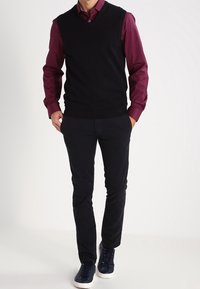 Pull sans manches noir porté sur une chemise bordeaux à manches longues, associé à un pantalon bleu marine et des baskets foncées, avec une texture lisse.