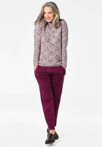 Grauer Strickpullover mit pinken Sprenkeln, kombiniert mit tiefpurpurfarbigen Wildlederhosen. Das Model steht mit den Händen in den Taschen und trägt schwarze Slipper.