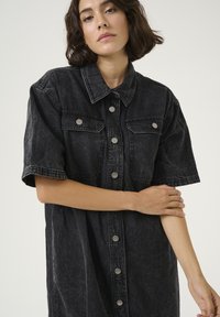 Vestido camisero de denim negro con mangas cortas, grandes bolsillos frontales y botones a presión plateados. Presenta un corte relajado y una textura desgastada.