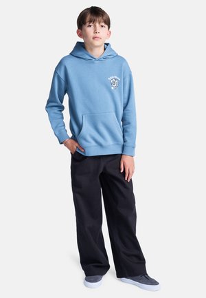 Lichtblauwe hoodie met een voorzak en logo-informatie op de linkerkant van de borst, gecombineerd met zwarte wijde broek en grijze schoenen.