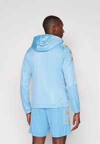 Hoodie bleu clair avec une capuche à cordon, arborant des accents de logo dorés sur les épaules. Accompagné de shorts bleu clair assortis. Texture lisse.