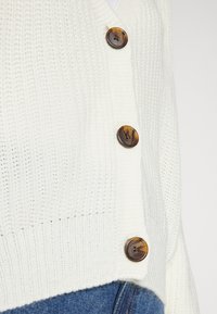 White knitted cardigan featuring a V-neck, four large, round brown buttons, and a textured pattern. Paired with blue denim.