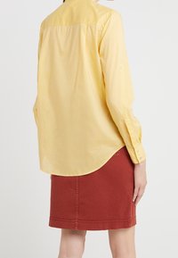 Camicia gialla a maniche lunghe con bottoni e orlo curvo, indossata sopra una gonna di jeans rossa. Il tessuto appare liscio con dettagli di cucitura visibili.