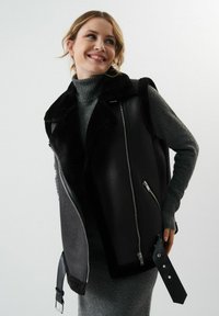 Czarna skórzana kamizelka z kołnierzem z sztucznego futra, z zamkami błyskawicznymi po bokach, detalem z klamrą oraz teksturowanym szarym golfem pod spodem.