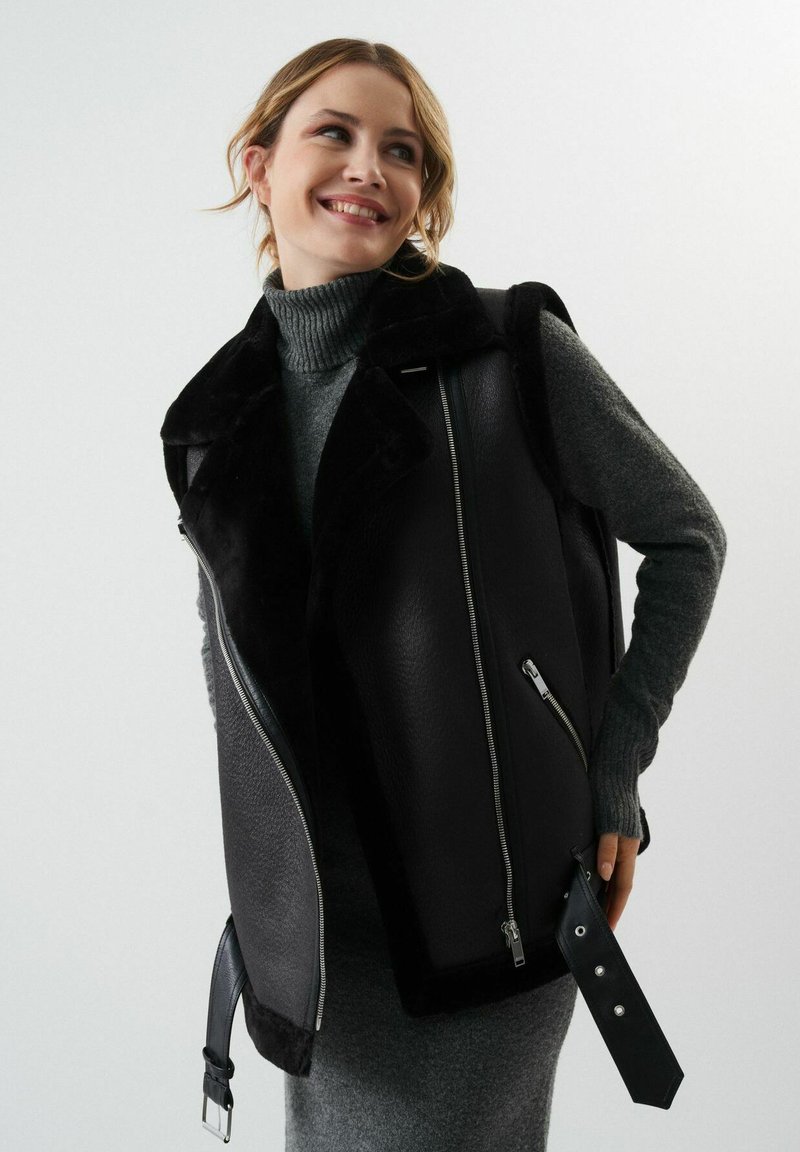 Czarna skórzana kamizelka z kołnierzem z sztucznego futra, z zamkami błyskawicznymi po bokach, detalem z klamrą oraz teksturowanym szarym golfem pod spodem.