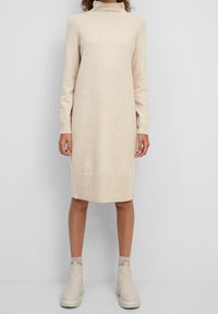Beiges Strickkleid mit hohem Kragen, langen Ärmeln und geripptem Saum. Getragen mit hellen Knöchelstiefeln und Stricksocken.
