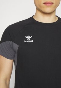 Homme portant un t-shirt noir et gris à manches courtes avec un logo hummel blanc sur la poitrine, montré des épaules au milieu du torse.