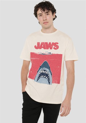 Cream-colored cotton t-shirt featuring a graphic of a shark with an open mouth against a red background, labeled "JAWS" in bold text.