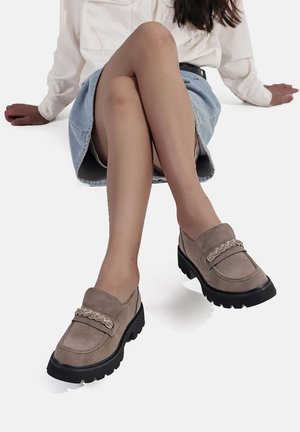 Mocassini in suede beige con una spessa suola in gomma nera e un'accorciatura decorativa in catena. Il design presenta una punta arrotondata e un tacco basso.