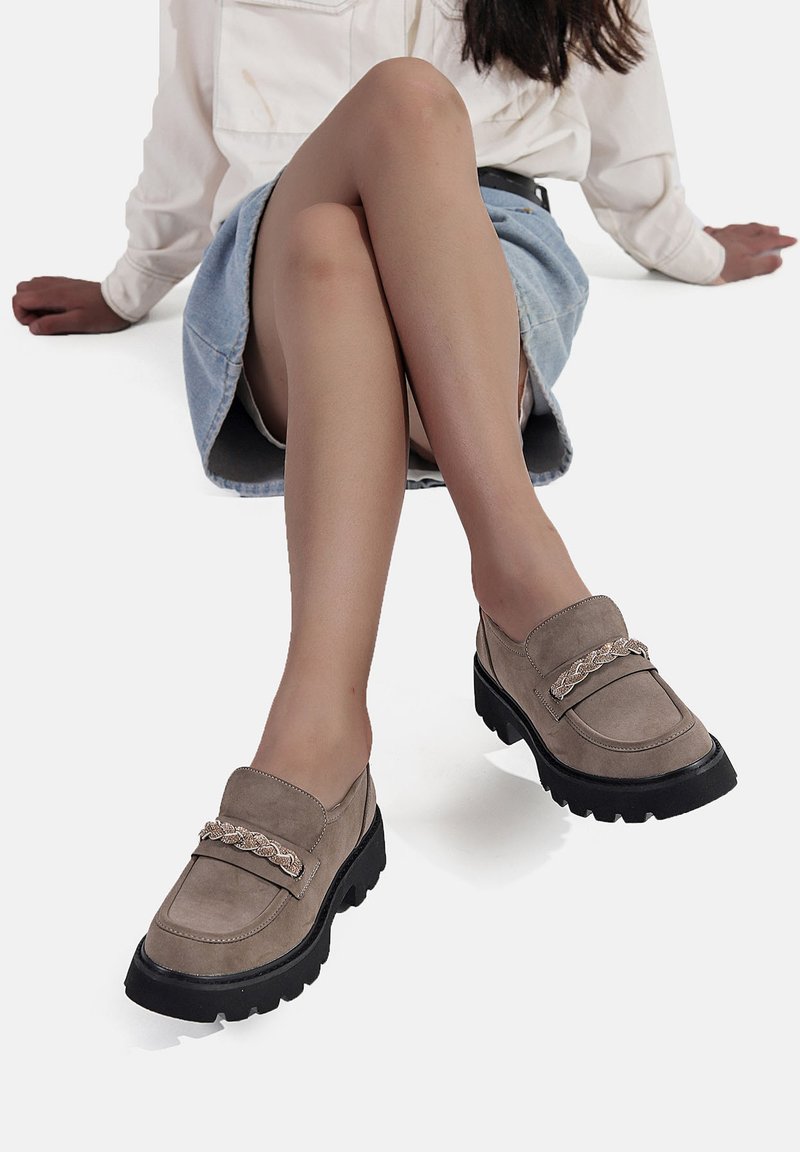 Mocassini in suede beige con una spessa suola in gomma nera e un'accorciatura decorativa in catena. Il design presenta una punta arrotondata e un tacco basso.