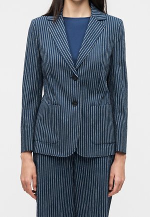 Kvinde iført en marineblå blazer med nålestriber med to sorte knapper og matchende nålestribede bukser, stående med armene afslappet ned langs siden.