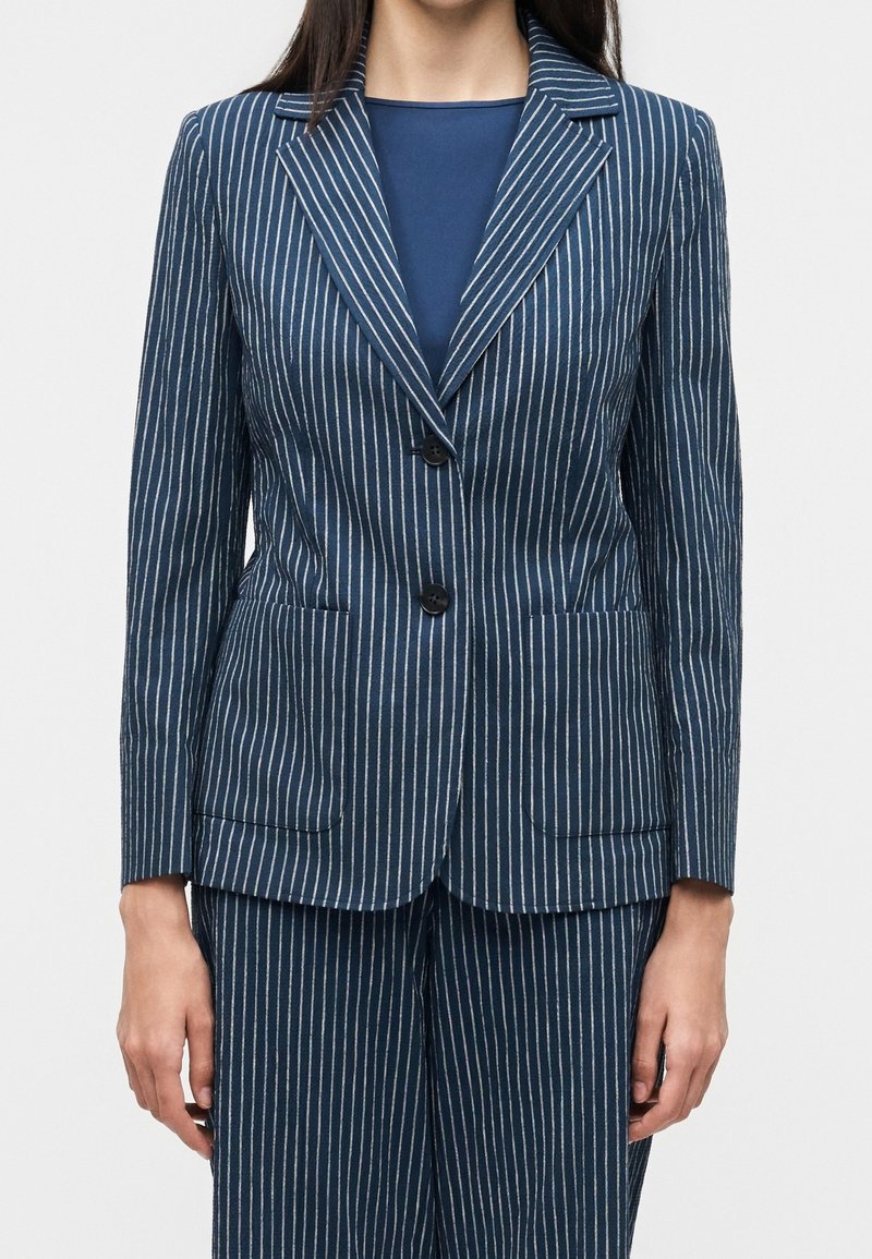 Femme portant un blazer bleu marine à fines rayures avec deux boutons noirs et un pantalon assorti à fines rayures, debout, les bras relâchés le long du corps.