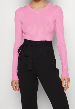 Femme portant un top à manches longues côtelé rose ajusté et un pantalon noir taille haute noué avec une ceinture en tissu à la taille.