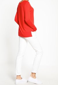 Person klädd i röd långärmad topp, vita uppvikta byxor och vita slip-on skor, stående i en avslappnad pose mot en vit bakgrund.