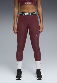 Burgunderfarvede atletiske leggings med en høj sort talje, der har hvid "PUMA" tekst. Glat tekstur, tætsiddende design, parret med sorte sneakers.