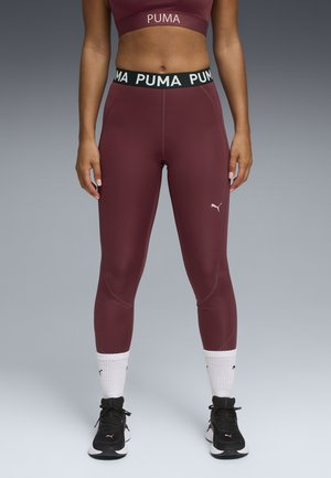 Burgunderfarvede atletiske leggings med en høj sort talje, der har hvid "PUMA" tekst. Glat tekstur, tætsiddende design, parret med sorte sneakers.