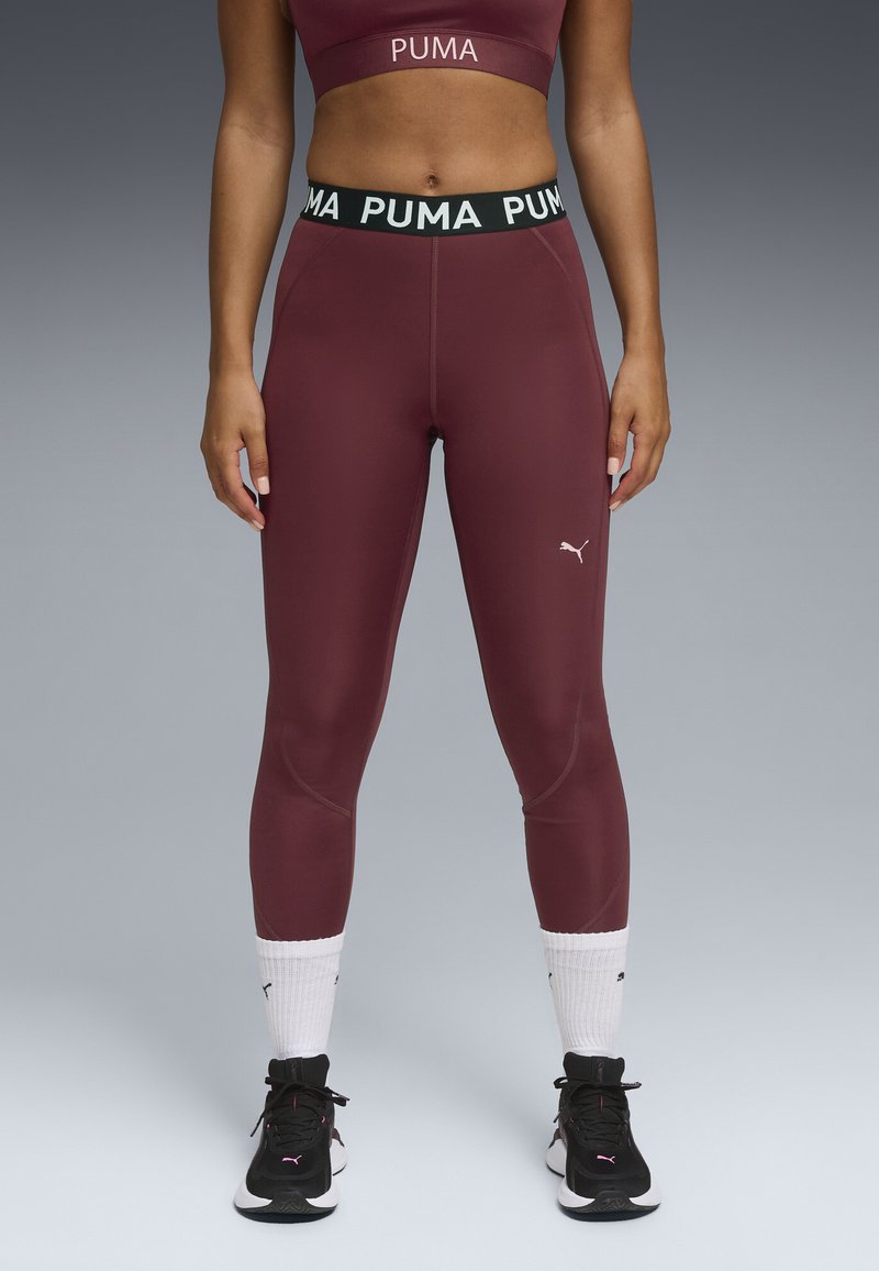 Burgunderfarvede atletiske leggings med en høj sort talje, der har hvid "PUMA" tekst. Glat tekstur, tætsiddende design, parret med sorte sneakers.