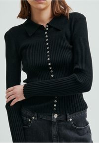 Svart ribbad stickad cardigan med krage, med metallknappar framtill och åtsittande ärmar. Bärs över mörka jeans.