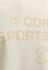 Tissu beige avec texte jaune délavé indiquant "EST. COR SPORTS" et un emblème circulaire de couronne de laurier avec des lettres à l'intérieur.