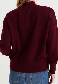 Burgundský pletený svetr s vysokým výstřihem, spadlými rameny a vroubkovanými manžetami, v kombinaci s světlými modrými džíny.