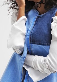 Gilet di denim con diverse sfumature di blu, dotato di chiusura con zip. Sotto, una maglietta bianca a maniche lunghe con dettagli in pizzo sulle maniche.