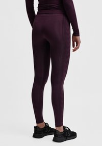 Leggings viola con una vestibilità aderente, motivi laterali tessurizzati e un tessuto liscio in contrasto. Abbinati a scarpe sportive nere.