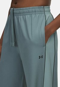 Light blue athletic pants made of smooth fabric, featuring an elastic waistband, side pockets, and a black logo on the thigh. Black piping details.
