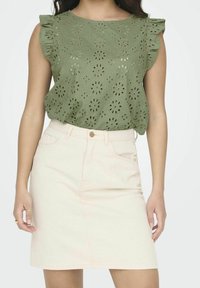 Blusa di cotone verde con ritagli floreali e maniche a volant, abbinata a una gonna di jeans rosa chiaro con tasche frontali e chiusura a bottone.