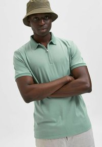 Světlezelená polo košile s krátkým zipem a síťovaným límcem. Model má na sobě béžový klobouk typu bucket, ruce zkřížené, na jednobarevném pozadí.