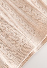 Texturerat stickat tyg i en mjuk beigefärg med en flätad mönstring och ribbade sektioner. Visar detaljerad sömnad och mjuk textur.