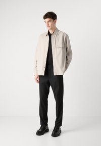Light beige button-up jacket with a chest pocket, black shirt underneath, black trousers, and black lace-up shoes. Smooth fabric texture.