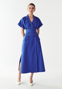 Mėlyna midi suknelė su dvigubais sagtais, trumpomis rankovėmis ir šoninėmis skeltukais. Pagaminta iš tekstūruotos medžiagos, turi dvi priekinės kišenes.