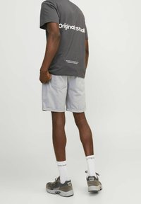 Camiseta gris con estampado "Original Studio", combinada con pantalones cortos gris claro con dobladillos enrollados, calcetines blancos y zapatillas grises con acabado texturizado.