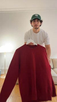 Mann trägt weißes T-Shirt und grüne Kappe, hält einen weißen Kleiderbügel mit einem großen roten Pullover in Innenräumen nahe einer Stehlampe und einem beigen Sofa.